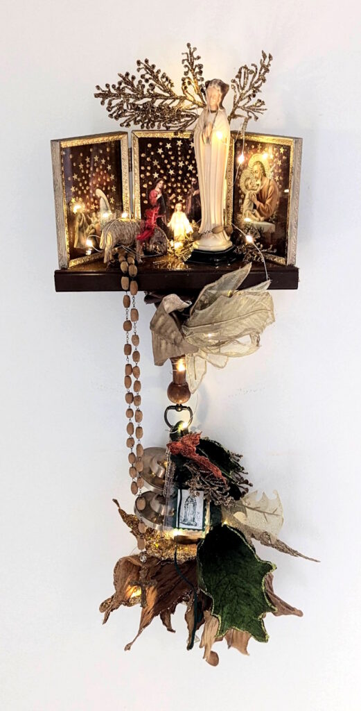 Alt text: Illuminated wall-mounted assemblage shrine featuring a central Virgin Mary figure, glowing lights, religious imagery, rosary beads, leaves, metallic branches, bells, and layered found objects against a white background.