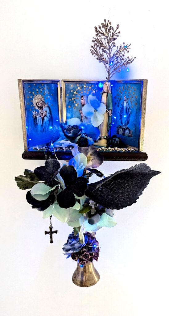 Illuminated wall-mounted assemblage shrine with Marian imagery, blue LED lights, flowers, a hanging cross, coral-like branches, and a brass bell against a white background.